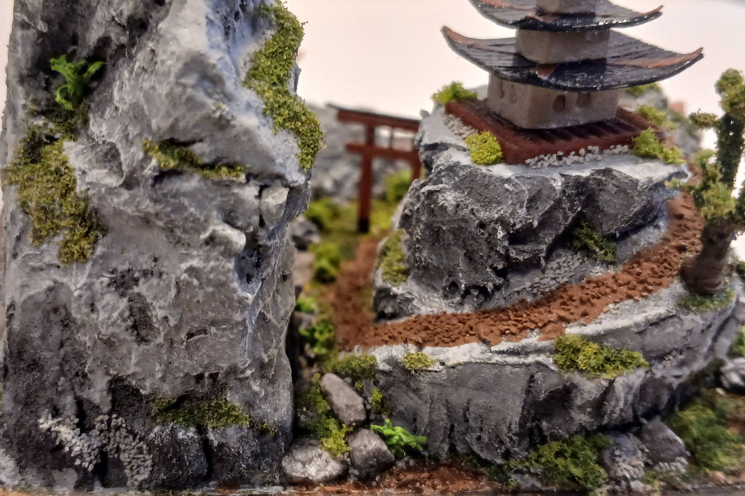 Miniature dirt path under a Torii Gate up a rock face mountain towards a shrine with cherry trees and foilage